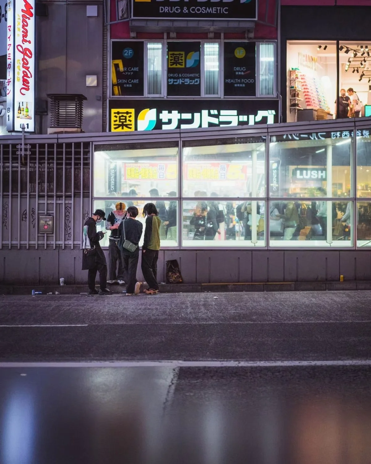 Shibuya nighttime city street scene with a group of five people standing and talking on the sidewalk in front of a glass-fronted building with bright signs, including a pharmacy and a store shot by Tokyo street photographer Deniz Demir.