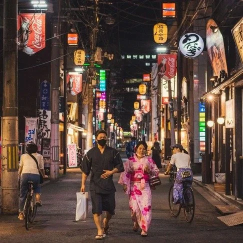 A couple in traditional Japanese attire walks down a lantern-lit Tokyo street at night, while cyclists and pedestrians move through the lively scene.