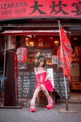 A cosplayer dressed in a pink outfit with oversized platform shoes and green hair sits in front of a traditional Japanese restaurant with red lanterns and flags. The sign above the restaurant is written in Japanese characters.
