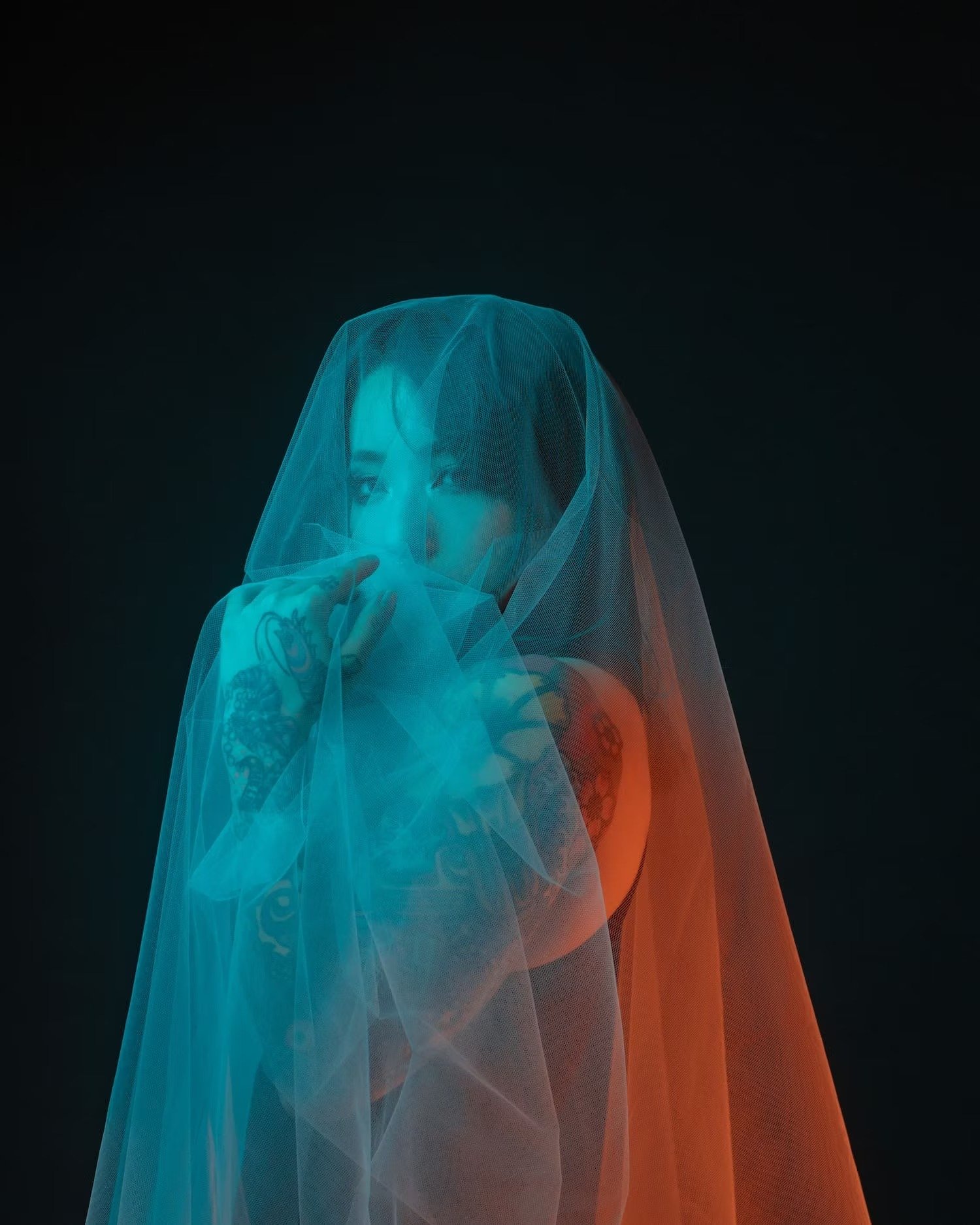 A person with tattoos is behind a semi-transparent orange and teal fabric, with a black background.