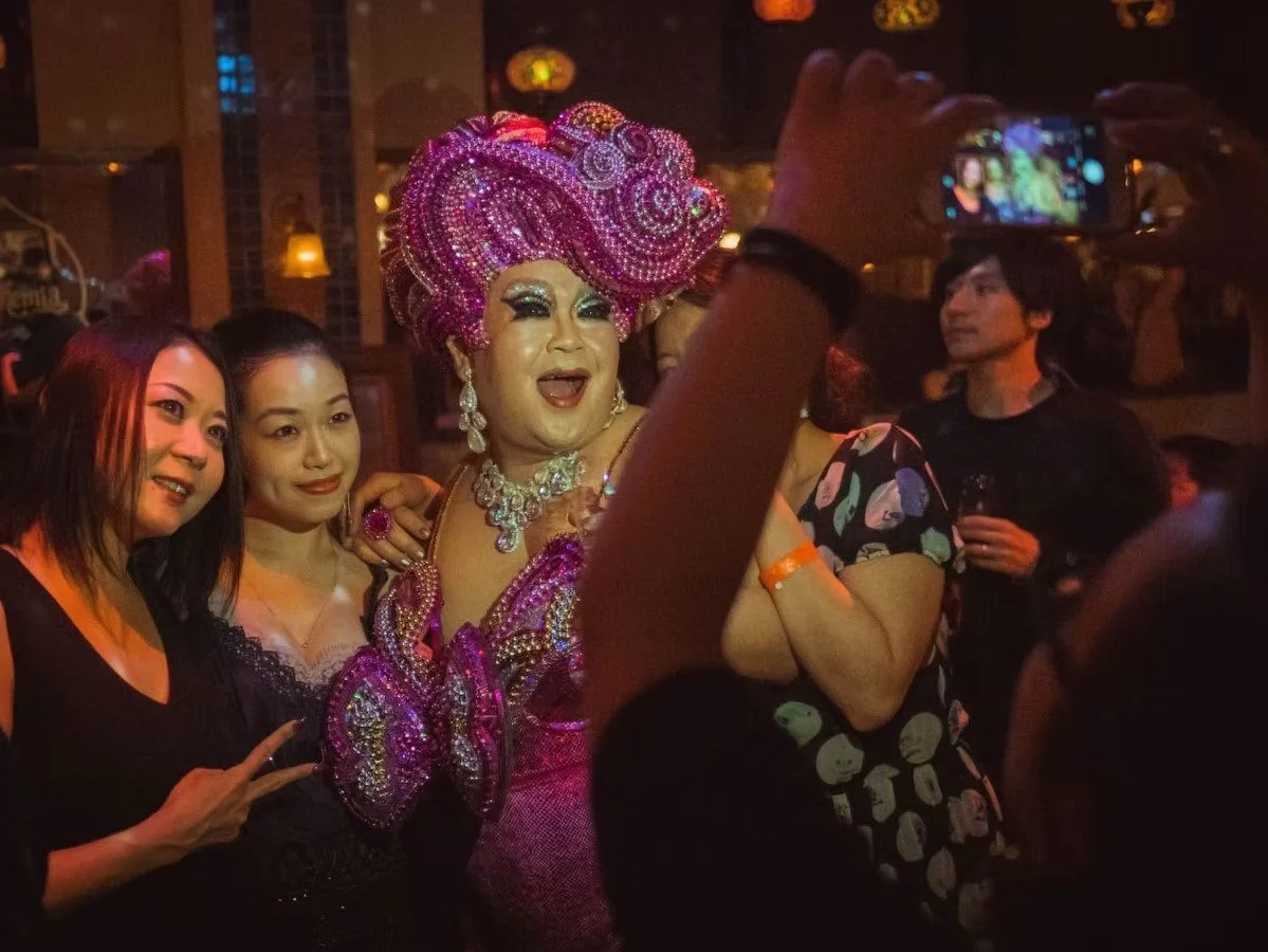 A drag queen with a large pink beaded headdress and matching pink outfit taking a selfie with three women at a dimly lit nightclub or bar. Night photography by Deniz Demir.