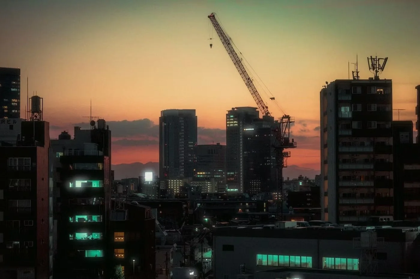 City skyline at sunset with tall buildings and a construction crane, illuminated windows, and a colorful sky in the background.