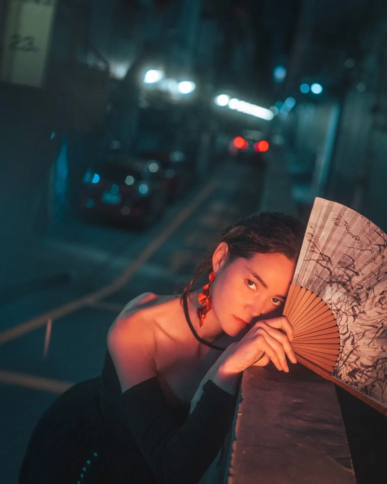 A woman with dark hair holding a decorative fan with a floral design, partially covering her face, posing in a dimly lit setting with a blurred background of streetlights and cars.