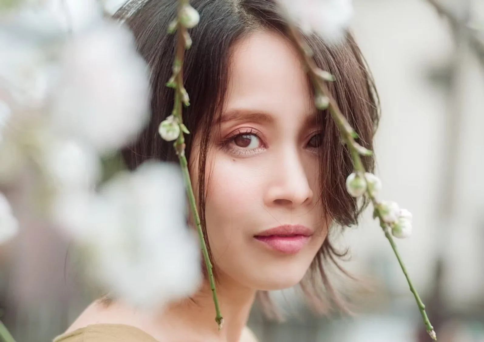A close-up portrait of a woman looking directly at the camera, framed by delicate cherry blossom branches. The soft focus of the flowers creates a dreamy, serene effect, highlighting her gentle expression.