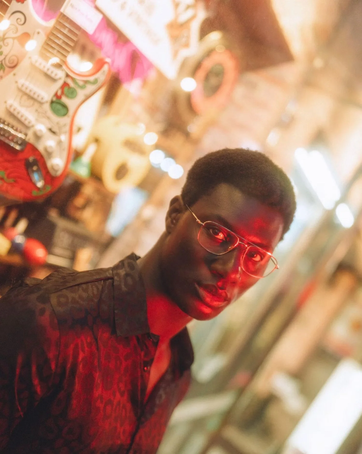 A person with dark skin and short hair wearing red-tinted glasses and a dark patterned jacket, surrounded by colorful blurred lights and decorations.