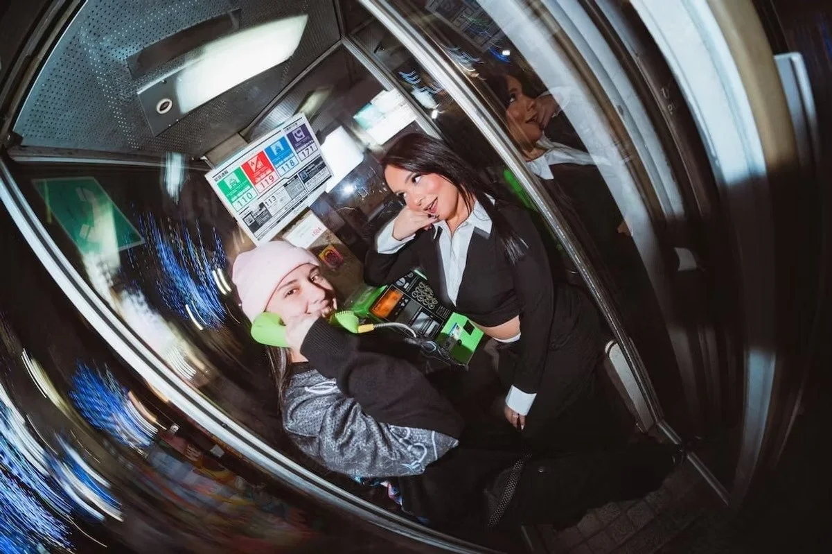 Fish eye photograph inside a glass phone booth showing two people posing playfully with a green telephone receiver. Reflections curve across the glass walls as city lights and signage distort around them, creating a nostalgic yet surreal urban scene.