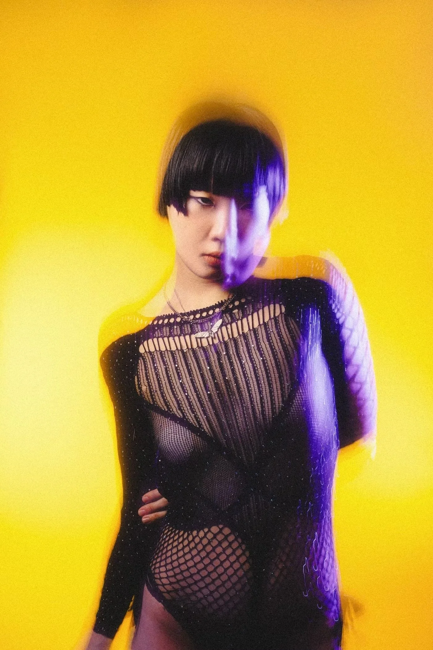 A person with short black hair and bangs, wearing a black fishnet dress with a silver necklace, standing against a yellow background, with purple lighting effects.