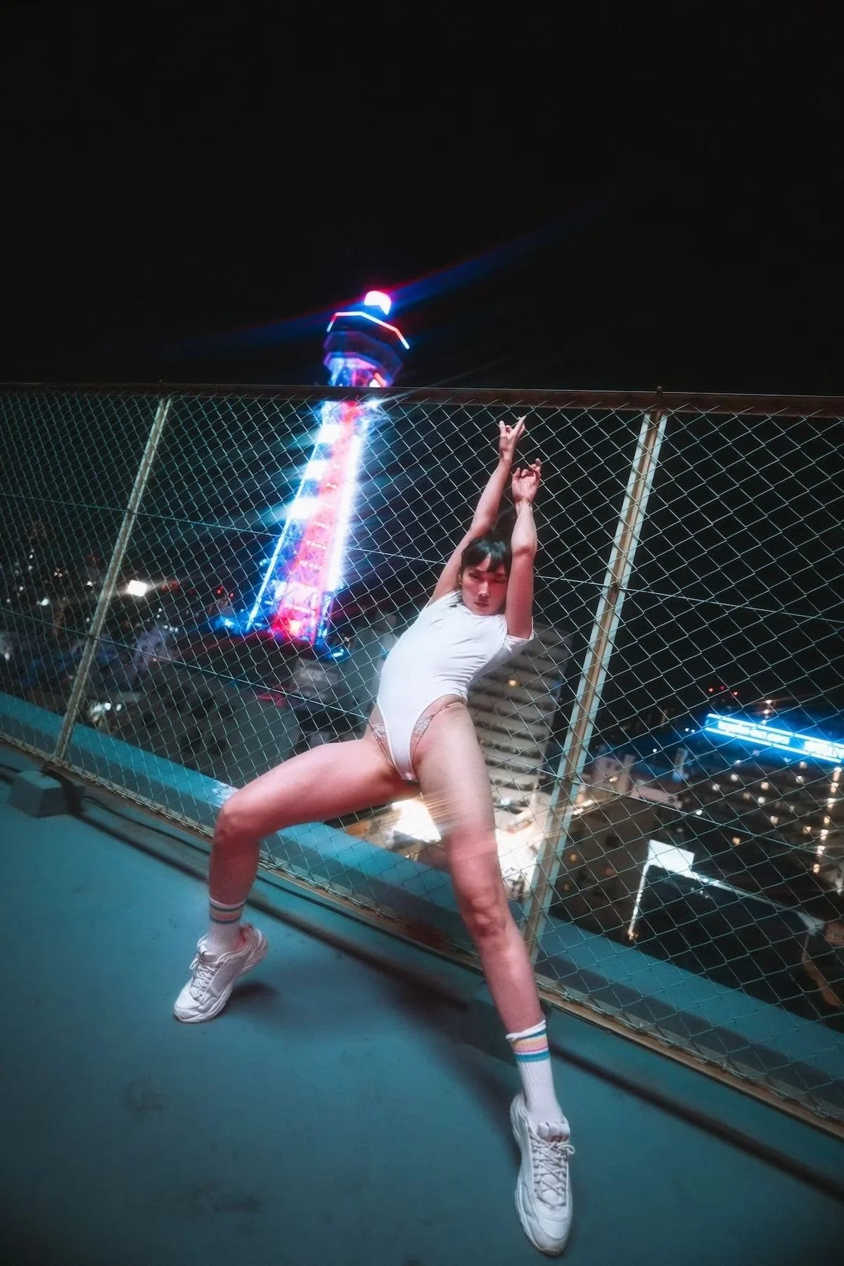A woman poses on a rooftop in Osaka at night with a cityscape and an amusement park ride with bright neon lights in the background. She is wearing a white outfit, sneakers, and colorful socks, striking a dance pose in front of a metal fence.