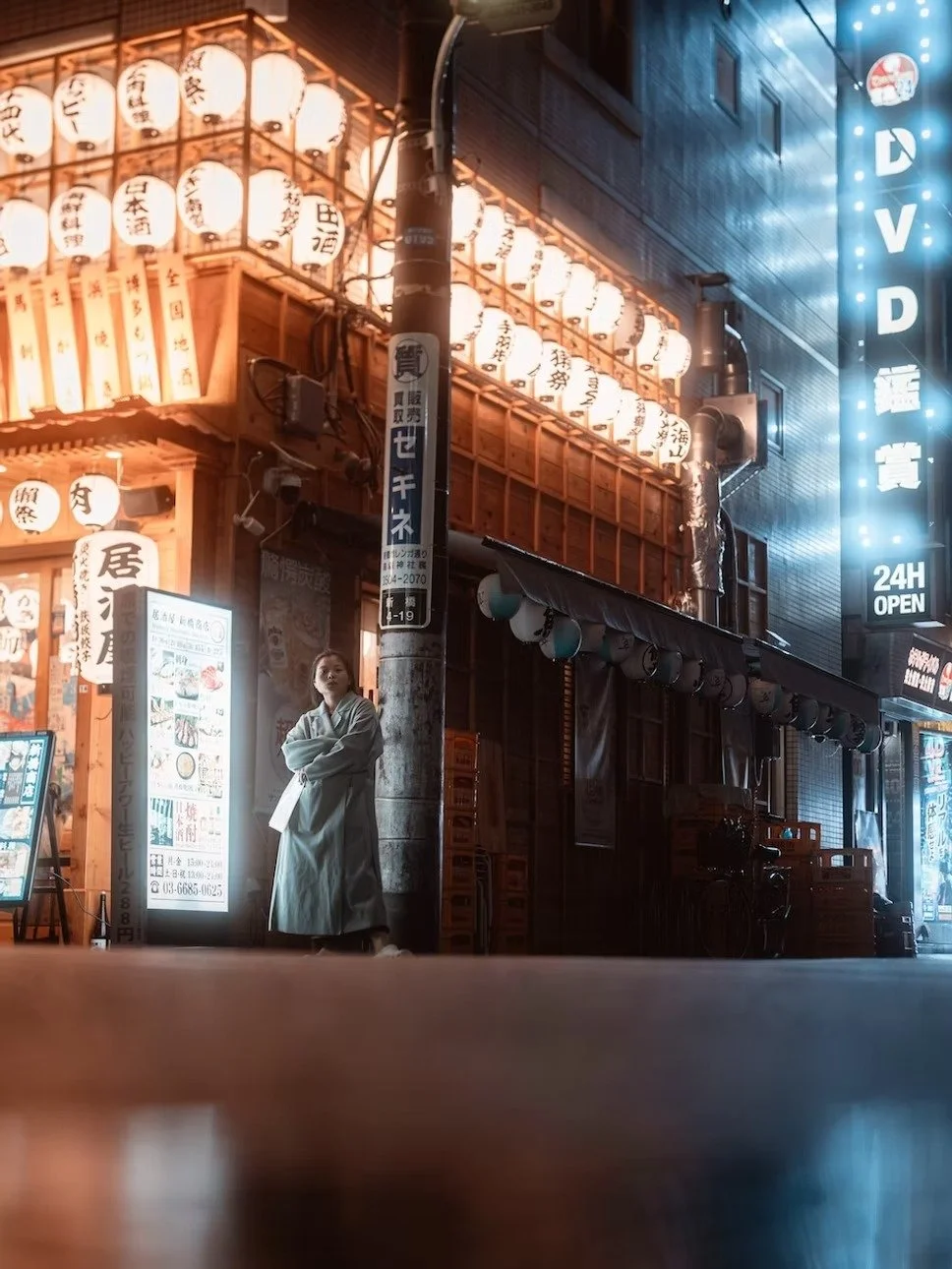 A woman standing outside a Japanese restaurant at night, illuminated by traditional paper lanterns with Japanese writing, with a neon sign indicating 24-hour service on a city street.