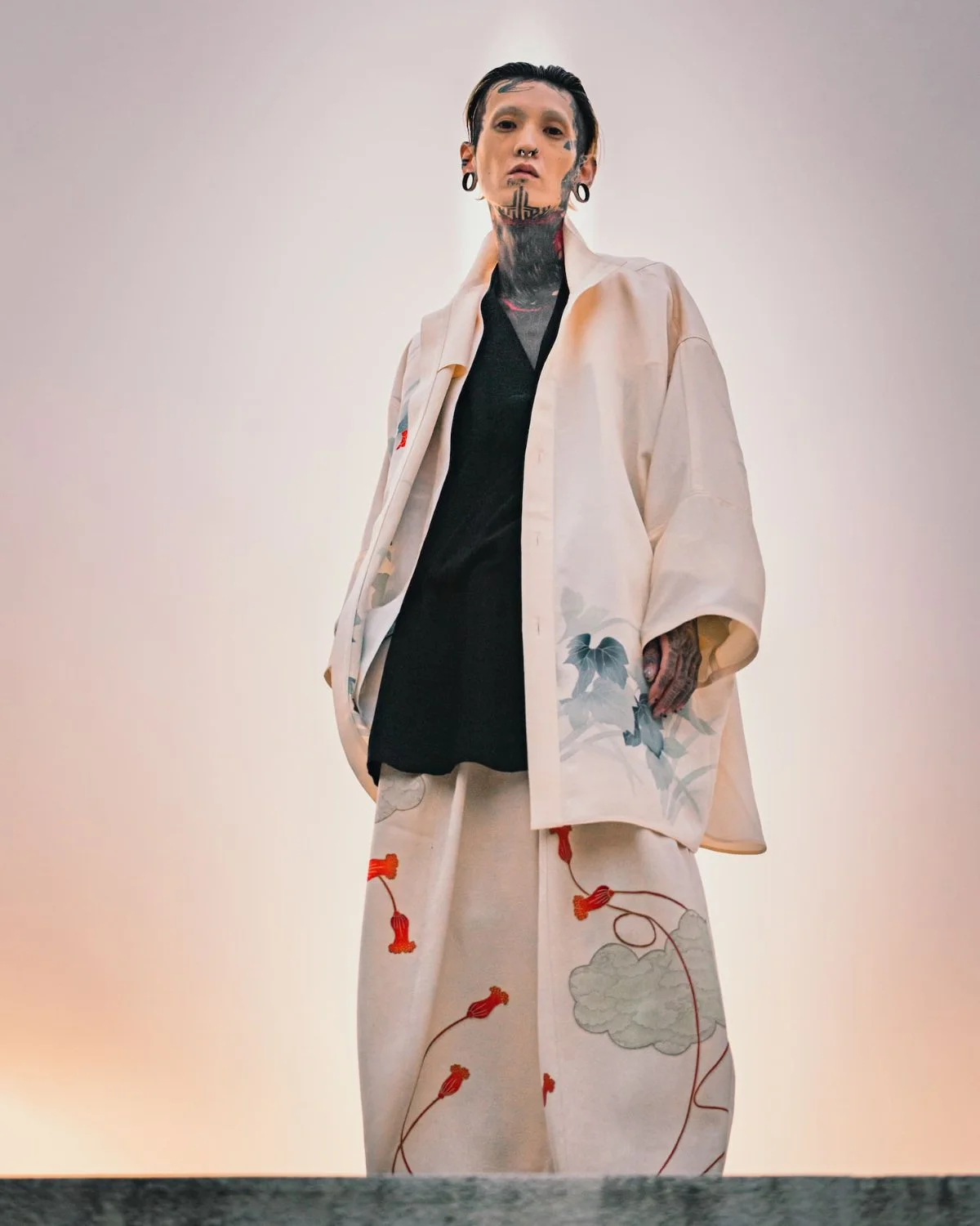 Man with tattoos and piercings wearing traditional Japanese-style kimono with cloud and floral pattern, standing against a plain background.