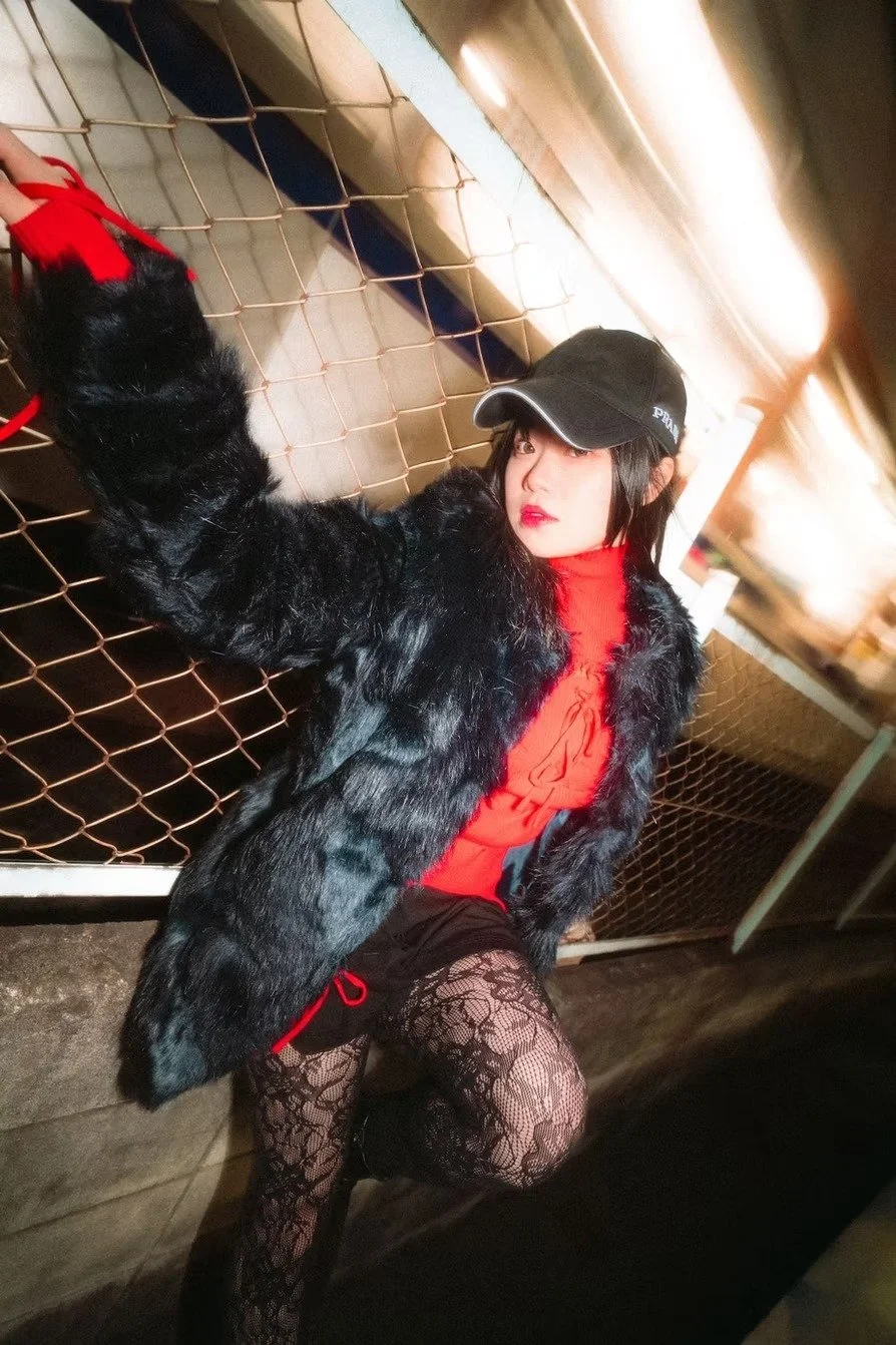 A woman with black hair wearing a black cap, red top, black fur coat, and lace tights, leaning against a chain-link fence shot by Tokyo portrait photographer Deniz Demir.