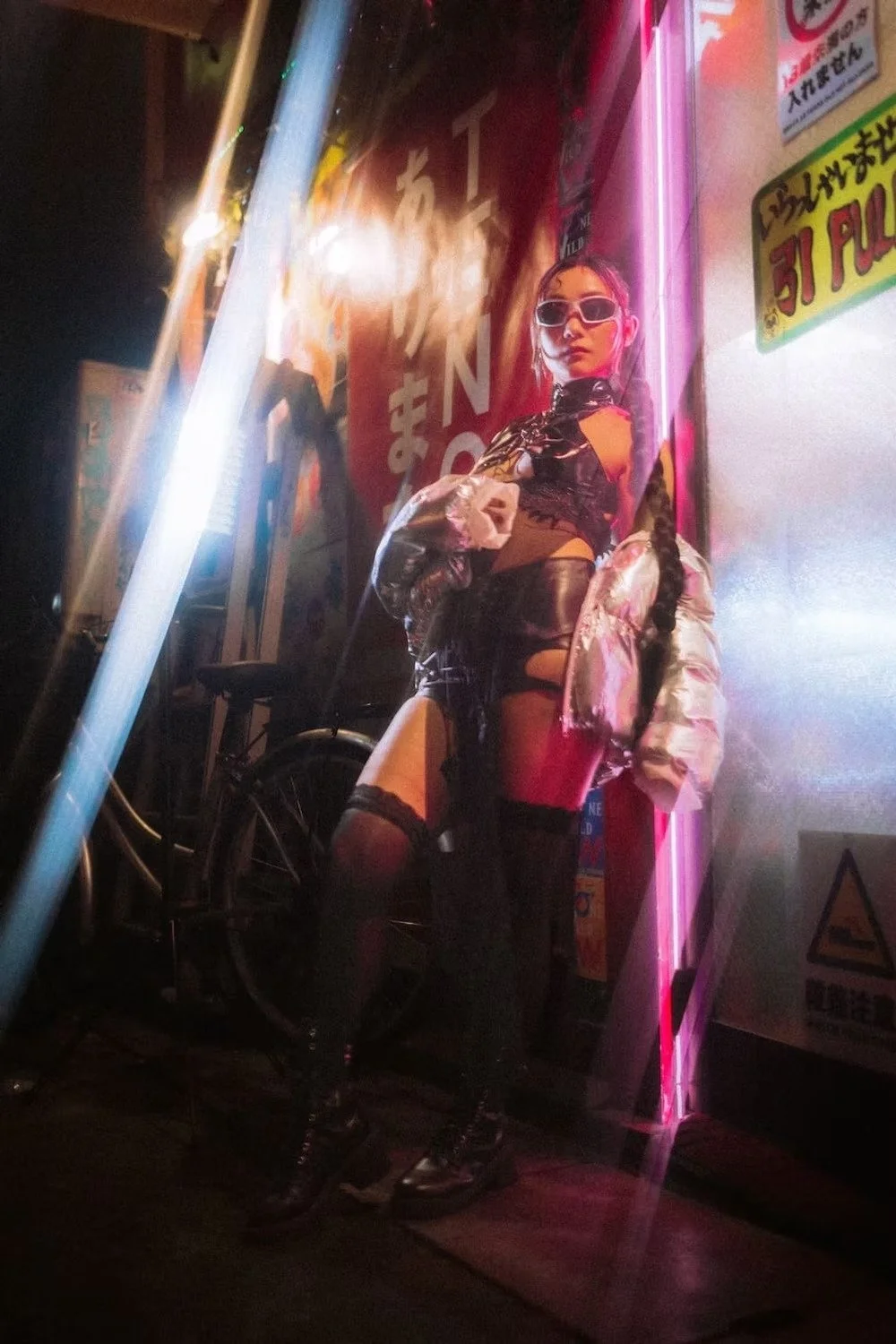 A woman dressed in black and metallic clothing with thigh-high stockings and sunglasses standing in a neon-lit street corner at night. She is leaning against a wall with neon signs and a bicycle nearby. Portrait photography by Deniz Demir.
