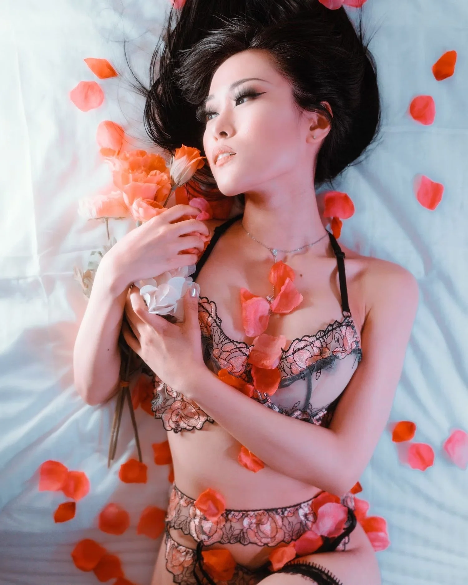 A woman with dark hair lying on a bed surrounded by scattered orange and pink rose petals, holding a rose near her face, and wearing lingerie with lace details and a necklace.