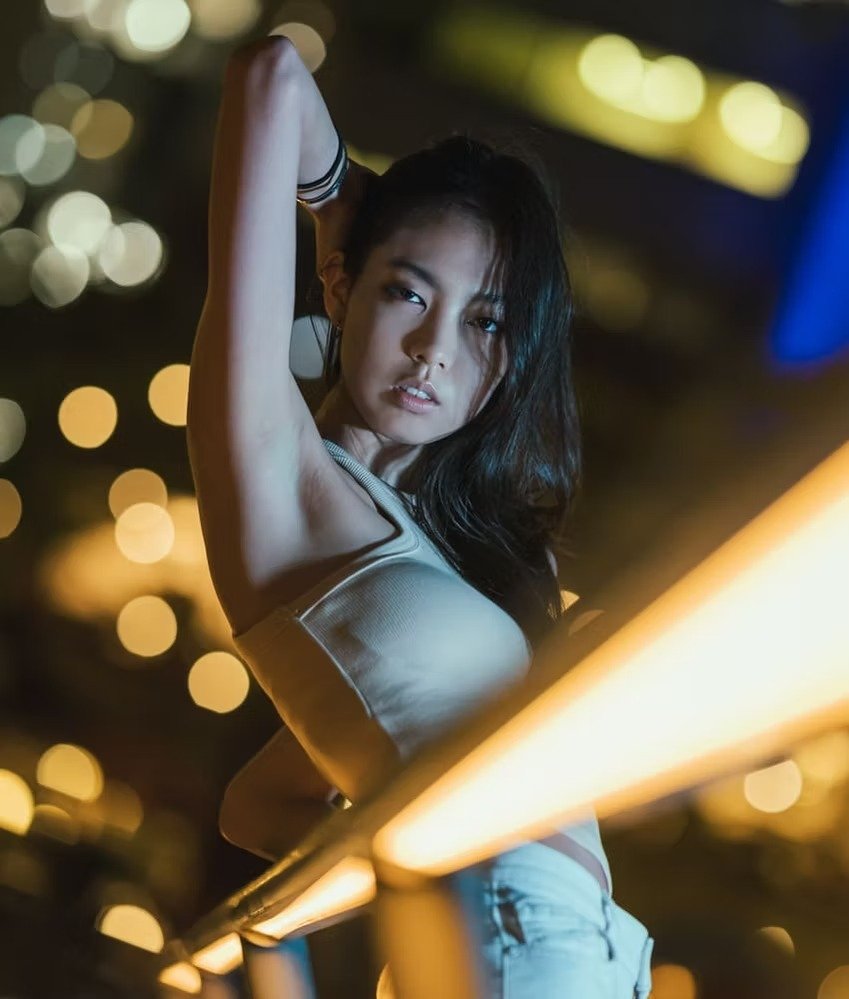 A young woman with dark hair posing at night with city lights in the background, wearing a sleeveless top and raising her arm behind her head.