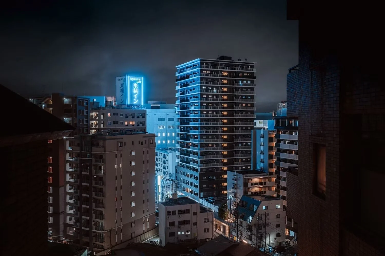 Nighttime cityscape view of tall buildings with illuminated windows and blue neon signs in an urban area.