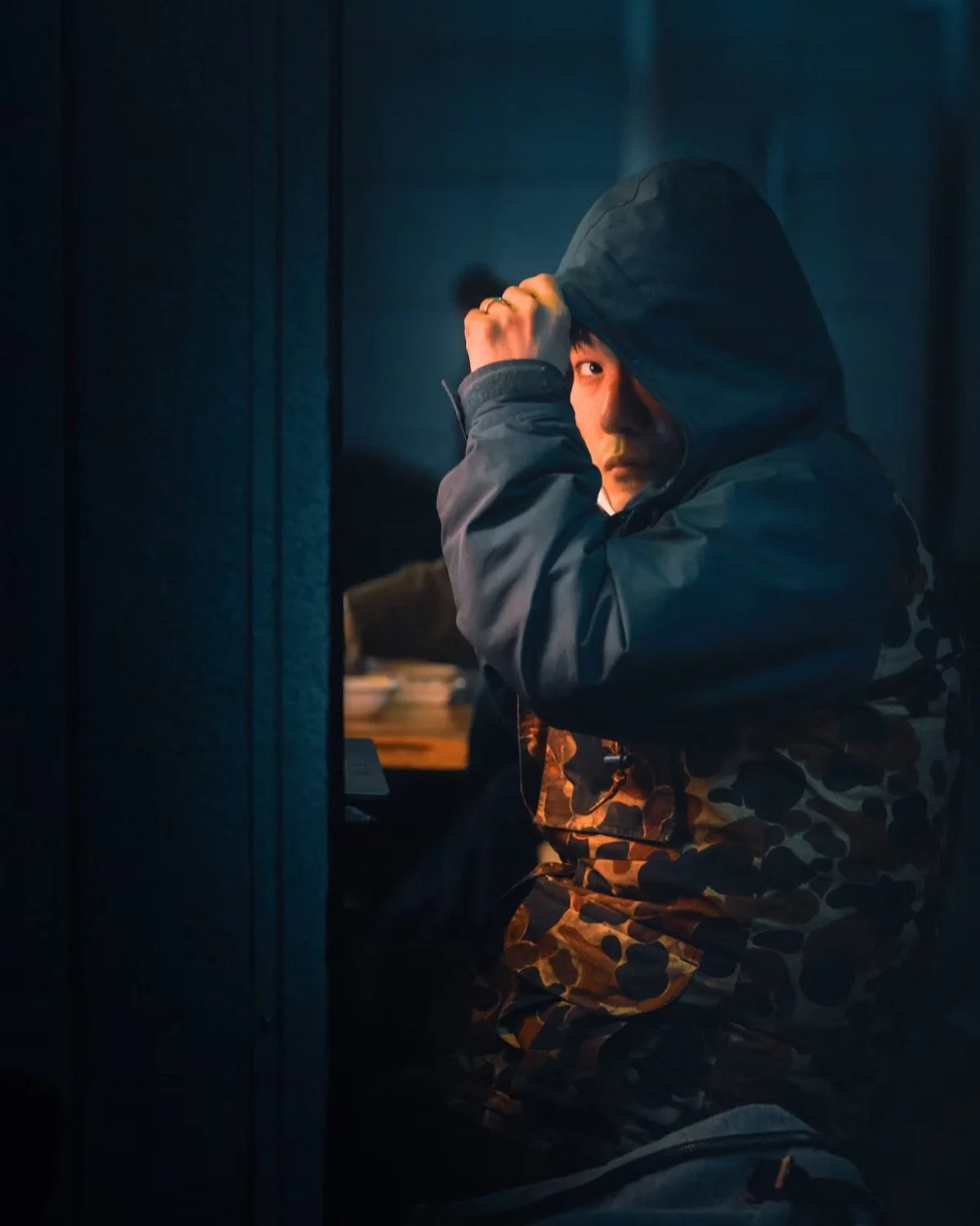 A young man in a camouflage jacket and hoodie, sitting inside a dimly lit room, looking over his shoulder shot by Tokyo street photographer Deniz Demir.