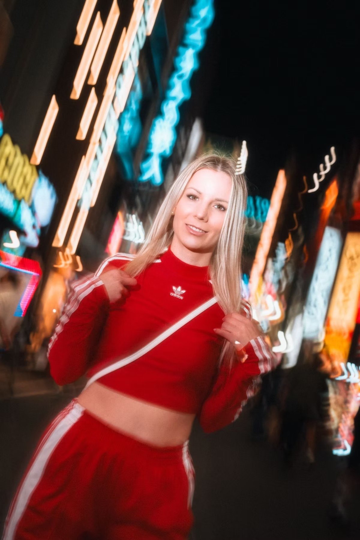 A woman in a red tracksuit stands confidently on a busy Tokyo street, illuminated by vibrant neon lights. The motion blur effect adds a sense of energy and movement. Street photography by Deniz Demir.