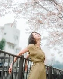Woman standing outdoors on a bridge near cherry blossom trees with pink flowers, looking up and smiling.