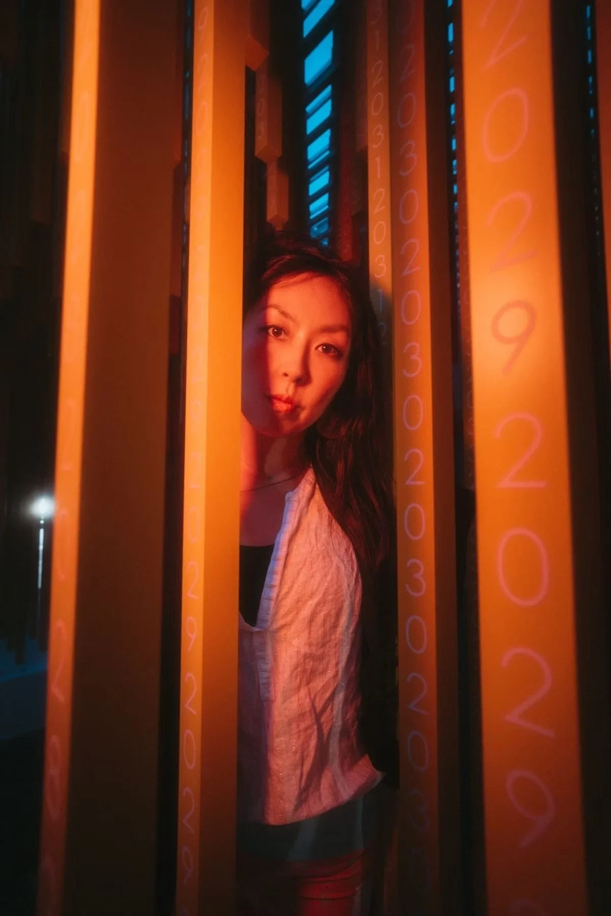 A Japanese woman with long dark hair looking through vertical orange bars with glowing blue and orange lights, numerical markings on the bars, in a dark setting.