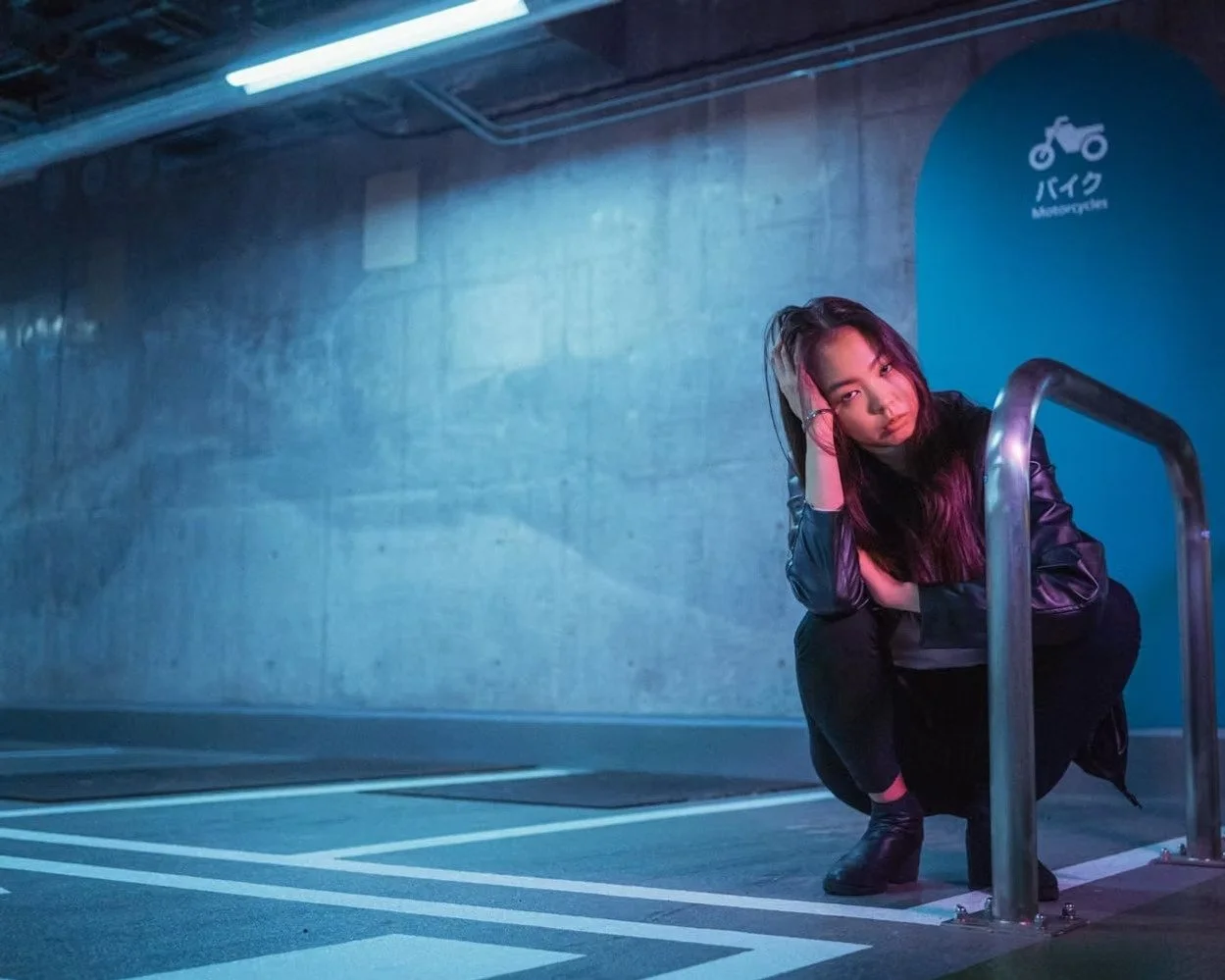 A young woman crouching near a bike rack in an underground parking lot, lit with blue and purple neon lights. She has long dark hair, wears a black jacket, and looks directly at the camera with a serious expression.