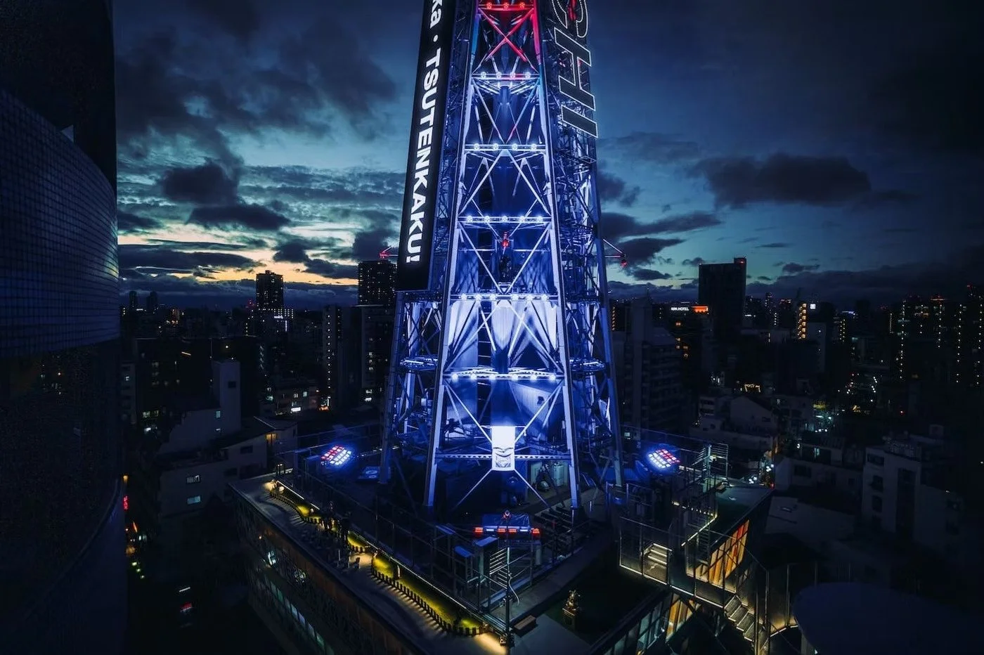 Nighttime cityscape featuring illuminated Tsutenkaku tower in Shinsekai Osaka with blue and purple lights, with surrounding buildings and clouds in the sky.