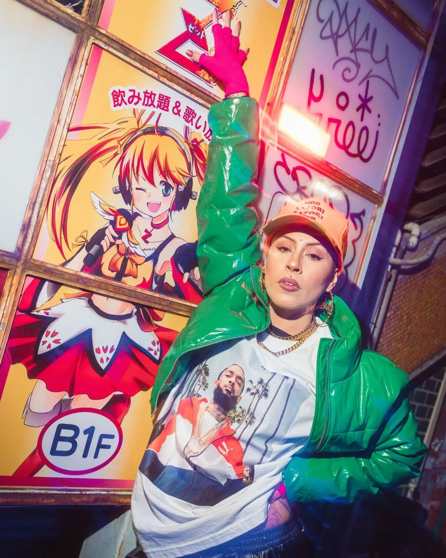 A person wearing a green shiny jacket, orange cap, background of colorful signs with Japanese writing, and a T-shirt with a man and palm trees. The person is making a peace sign.