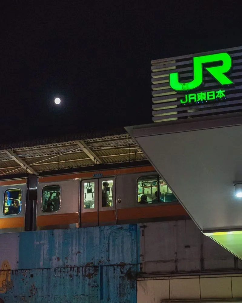 Night scene with a train partially visible on the platform, a bright moon in the dark sky, and a large illuminated green JR sign indicating a train station in Japan.