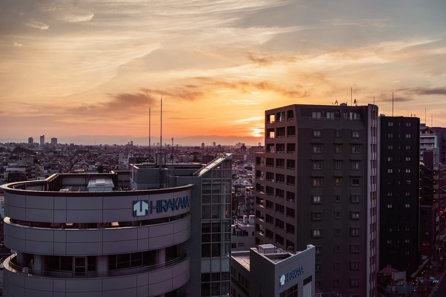 Sunset over a city skyline with tall buildings, including a curved building with the HRAKANA logo, in the foreground.