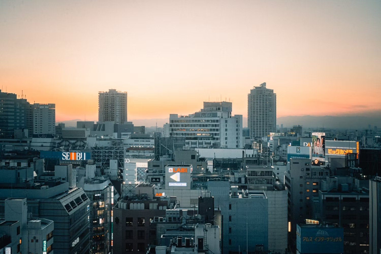 City skyline during sunset with tall buildings and illuminated signs in Japanese and English.