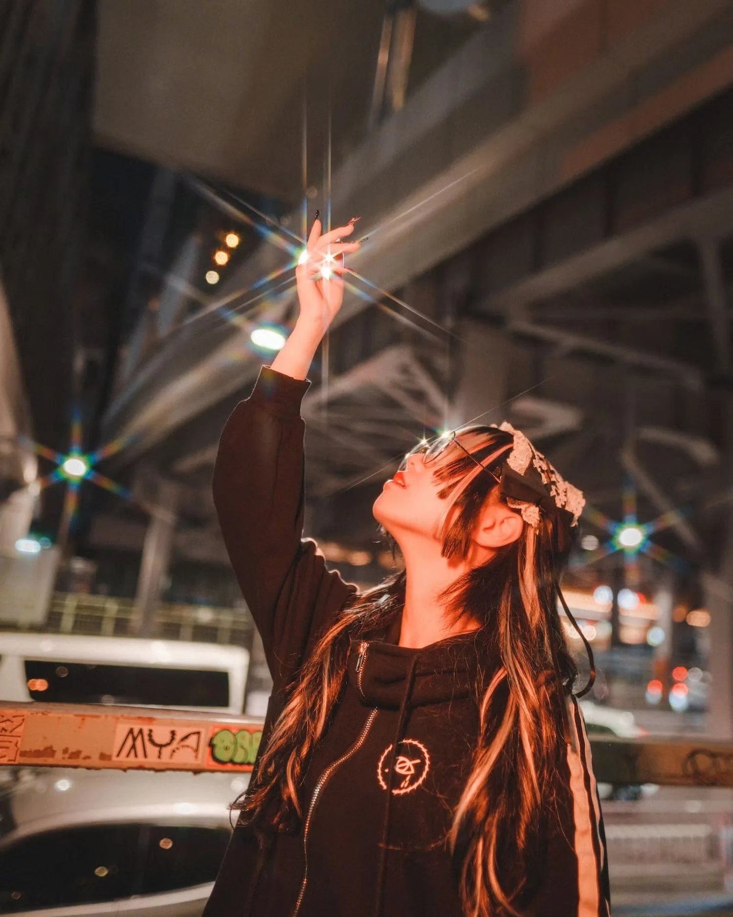 A woman with long dark hair and glasses, wearing a black jacket, is reaching up with her hand, and a bright light or flare is just above her fingers. The background appears to be an urban setting at night, with blurred lights and signs.