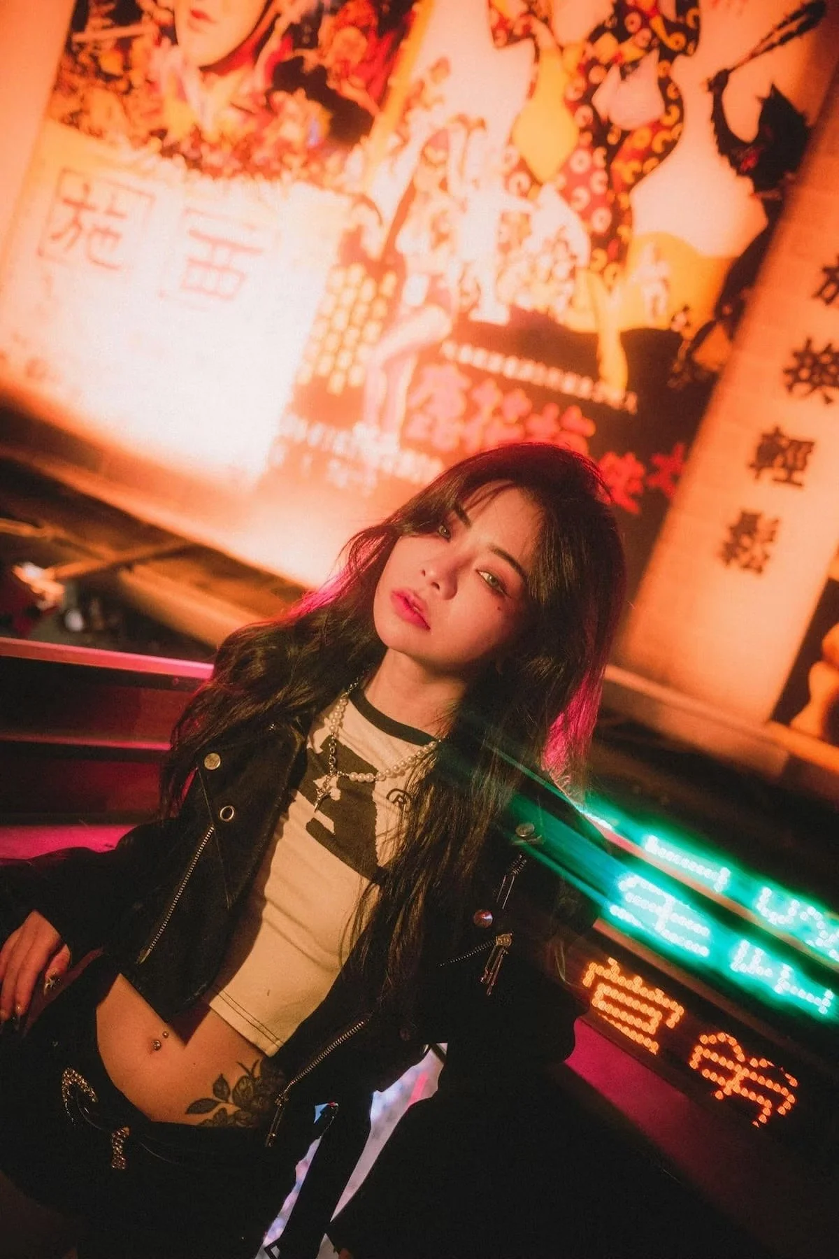 A young Taiwanese woman in with long black hair and makeup, wearing a black leather jacket, a beige crop top, and multiple necklaces, posing in front of a colorful neon-lit background with Asian-style banners and signs.