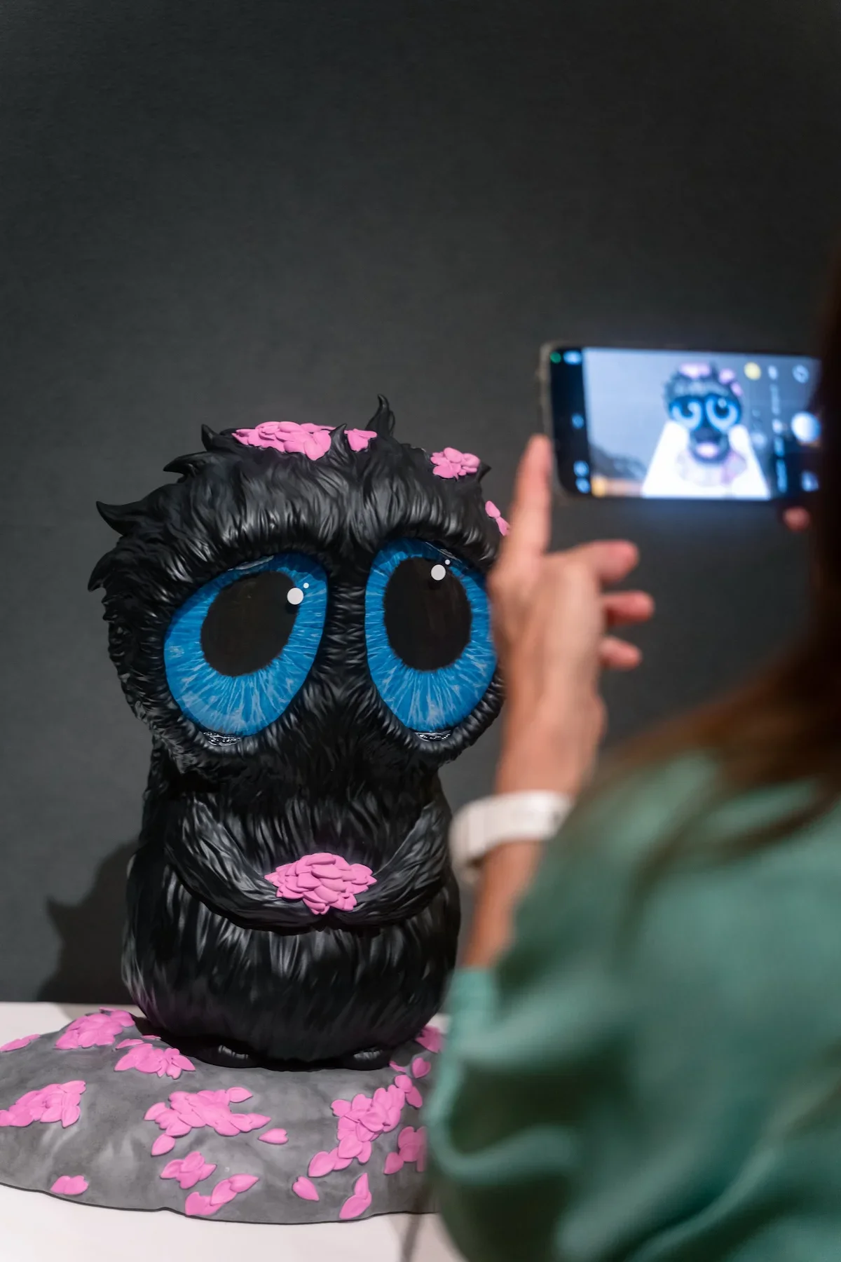 August Vilella
---
Close-up event photograph of a whimsical black creature sculpture featuring oversized blue eyes and pink decorative details, photographed as a visitor captures the artwork on a smartphone during August Vilella's exhibition.