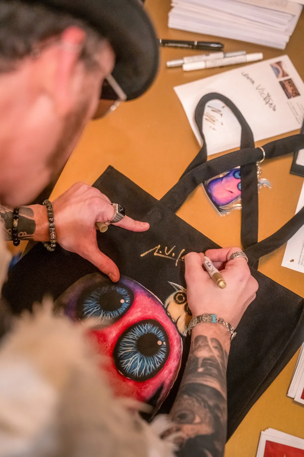 August Vilella
---
Detailed overhead photograph of August Vilella's tattooed hands signing a colourful surrealist art print featuring one of his signature wide-eyed creatures. A candid moment documenting the artist's interaction with collectors durin