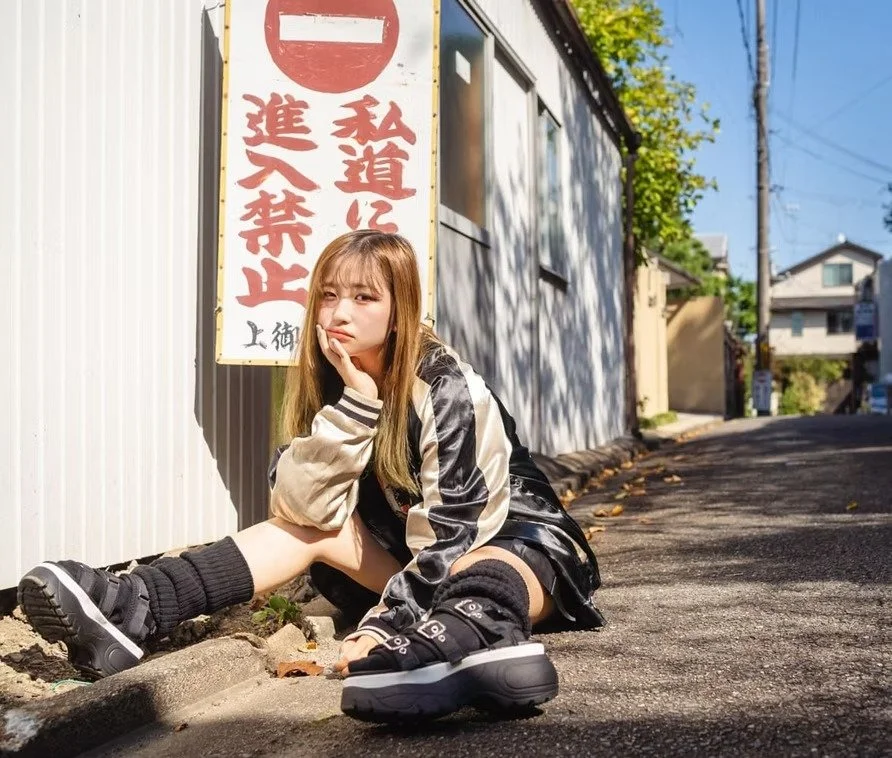Young woman with long hair sitting on the pavement, resting her chin on her hand, next to a sign with Japanese characters, in a sunny outdoor setting with buildings and trees in the background.
