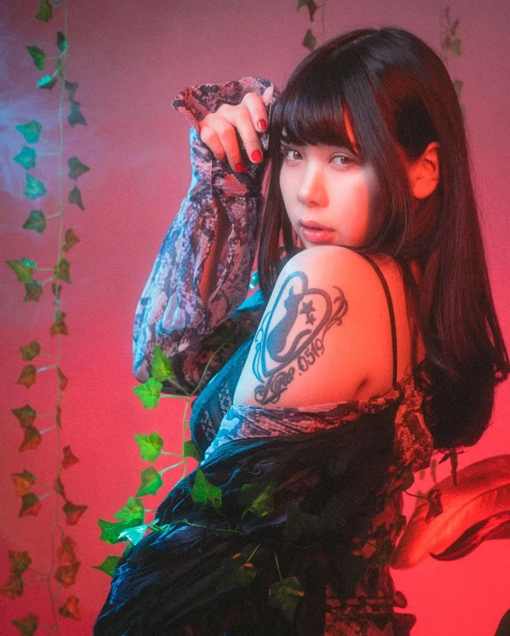 A woman with tattoos poses with a fierce expression against a vibrant red background. Her posture and intense gaze convey strength and attitude.