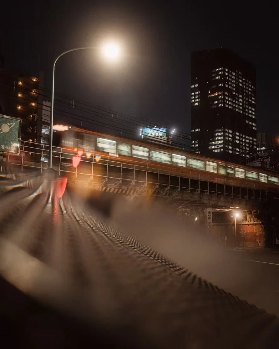 Nighttime Tokyo city scene with a moving train, illuminated from inside, passing above on an elevated track. Bright moon in the sky, large dark building with lit windows in the background, and a streetlamp casting light on the scene.