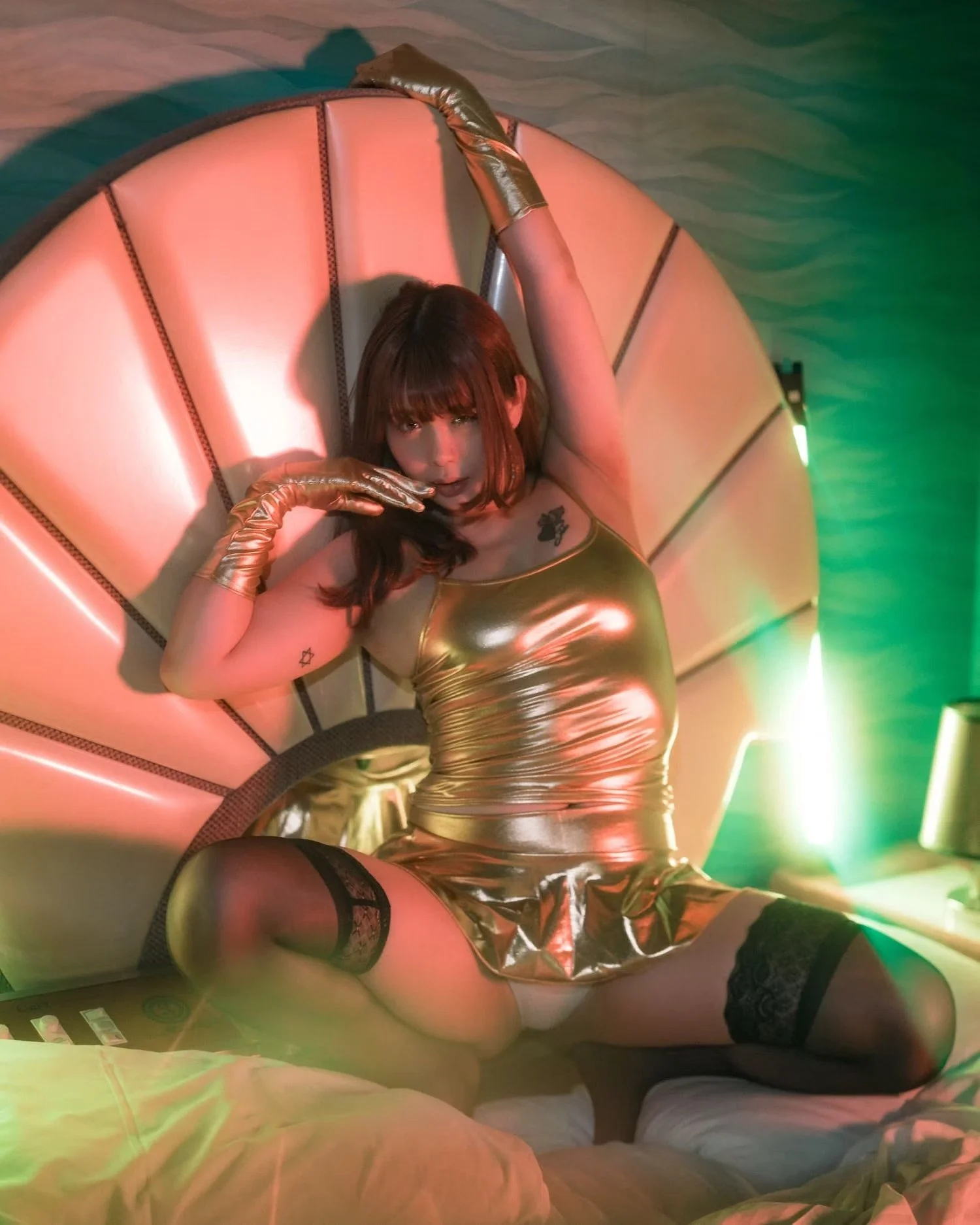A woman with brown hair, wearing gold clothing, thigh-high stockings, and gloves, laying on a large, illuminated, padded circular bed with a colorful, patterned backdrop.