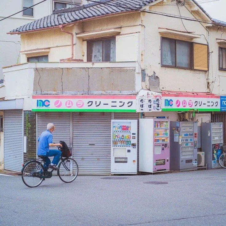 An elderly man riding a bicycle past vending machines in Osaka, captured during slow travel in the city.