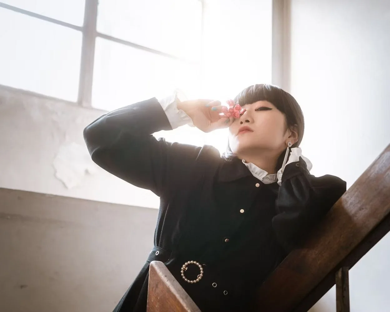 A Japanese girl in a black jacket and white blouse sitting on a wooden bench, holding a pink flower to her nose with her eyes closed in a bright room with large windows.
