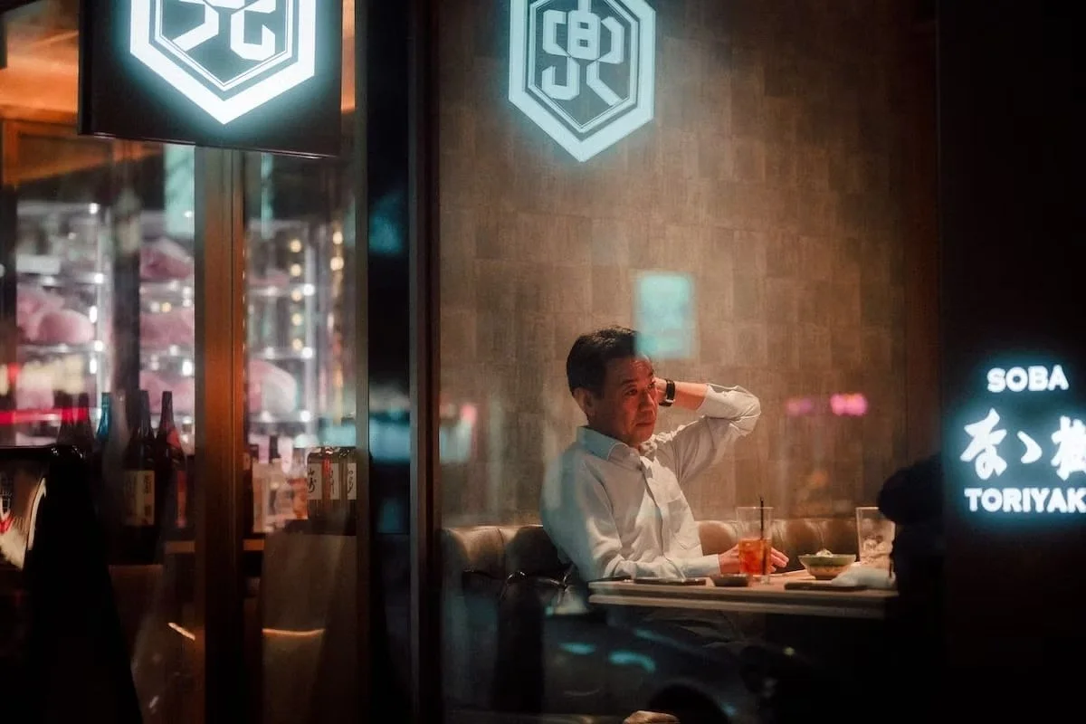 A man sitting at a restaurant table with a drink and food, adjusting his hair or head, inside a dimly lit Japanese restaurant with bottles on display. Yurakucho in Tokyo.
