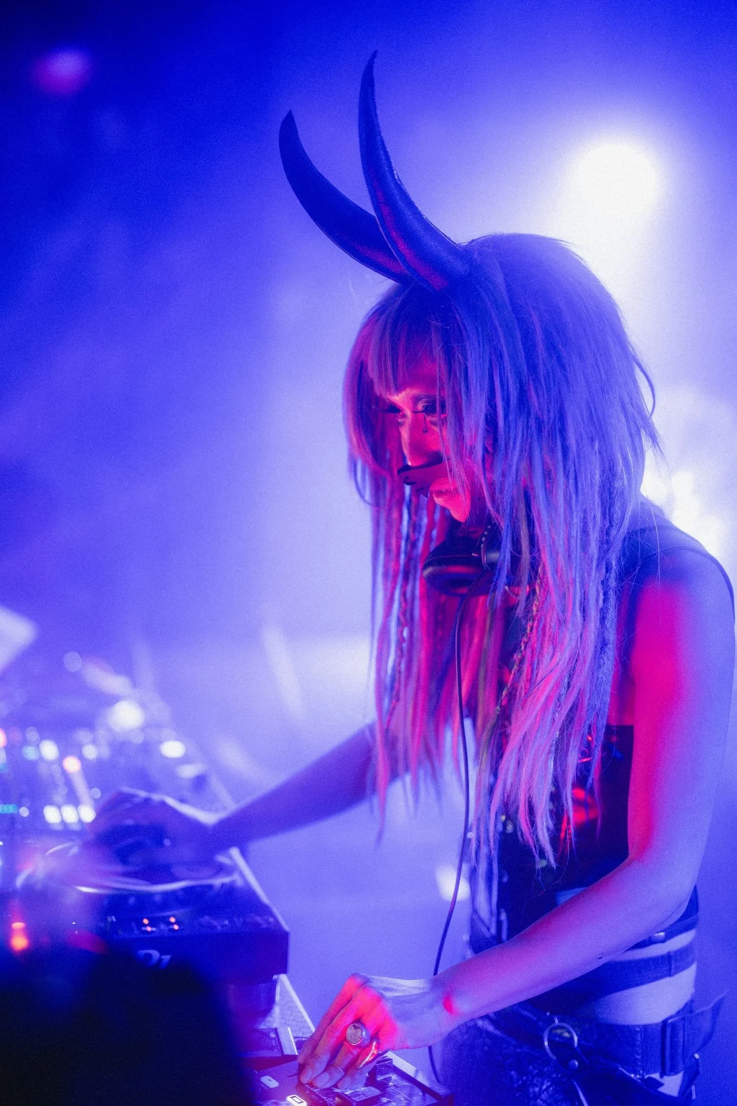 Bondage Ball 
---
A female DJ with long, colorful, wavy hair wearing glasses and a black tank top, playing music at a club or concert with blue and purple lighting and fog effects.
