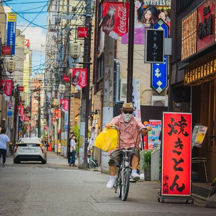 A man wearing a mask and sunglasses rides a bicycle on a narrow Tokyo street, surrounded by red lanterns, Japanese signs, and urban storefronts.