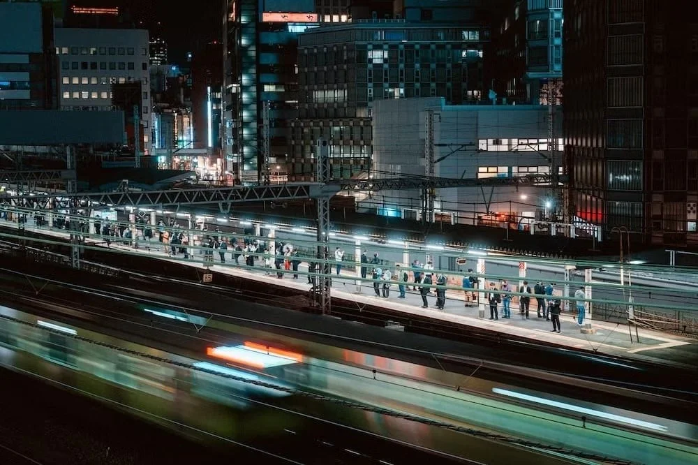 Nighttime scene of a busy city Shimbashi train station platform with people waiting in line, surrounded by tall illuminated buildings and blurred moving vehicles on the roads.