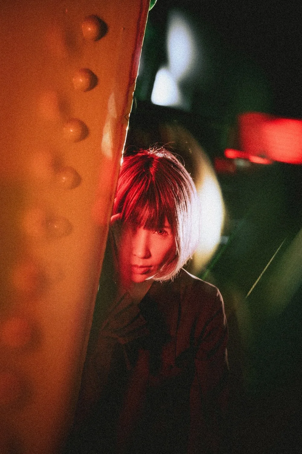 A Japanese woman with short hair, illuminated by red and white lights, peeking from behind a large, textured orange pillar in a dark setting with blurred colorful lights in the background shot in Shibuya by Tokyo portrait photographer Deniz Demir.