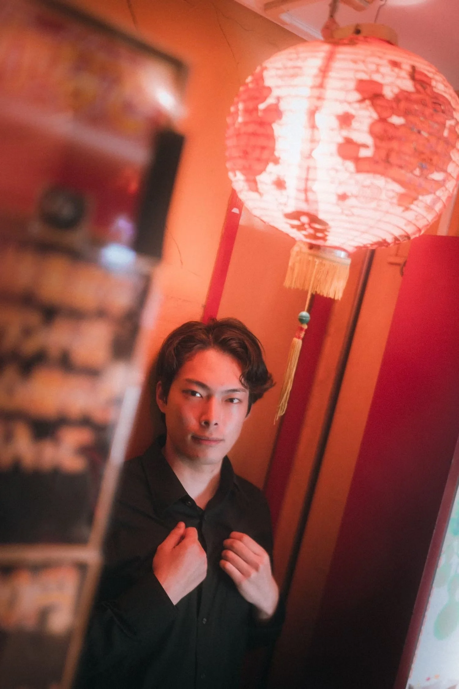 A young man with dark hair and a black shirt is sitting in a dimly lit room, with a red Chinese paper lantern hanging in the background. The photo is taken through a blurred object in the foreground.