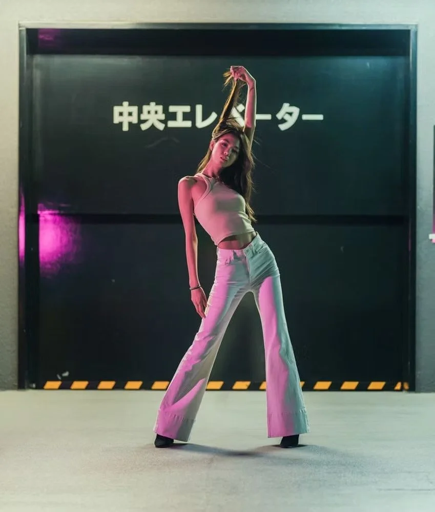 A woman in white crop top and wide-leg jeans poses with one arm raised above her head in front of a black door with Japanese characters.