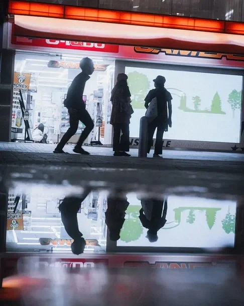 The silhouette of people walking along a rain-soaked street, with reflections of glowing billboards in the water. A perfect scene for street photography during Tekumi Photowalks.