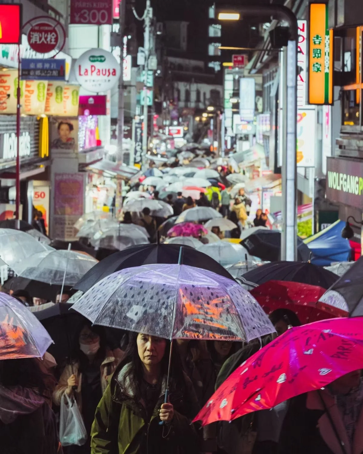 A crowded Harajuku Tokyo street during rain with many people holding umbrellas, neon signs, and buildings lining the street