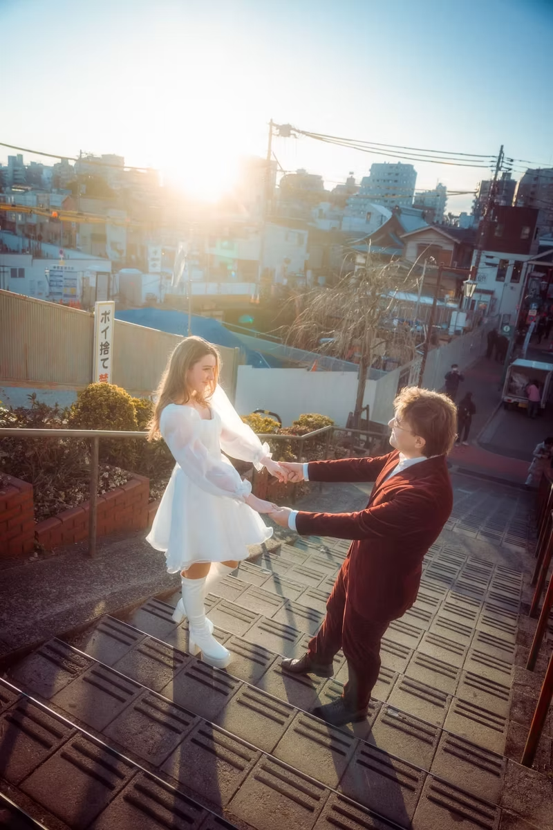 A couple dressed in white winter clothing holds hands while walking through a park in Tokyo, with sunlight streaming through the trees and the cityscape in the background.