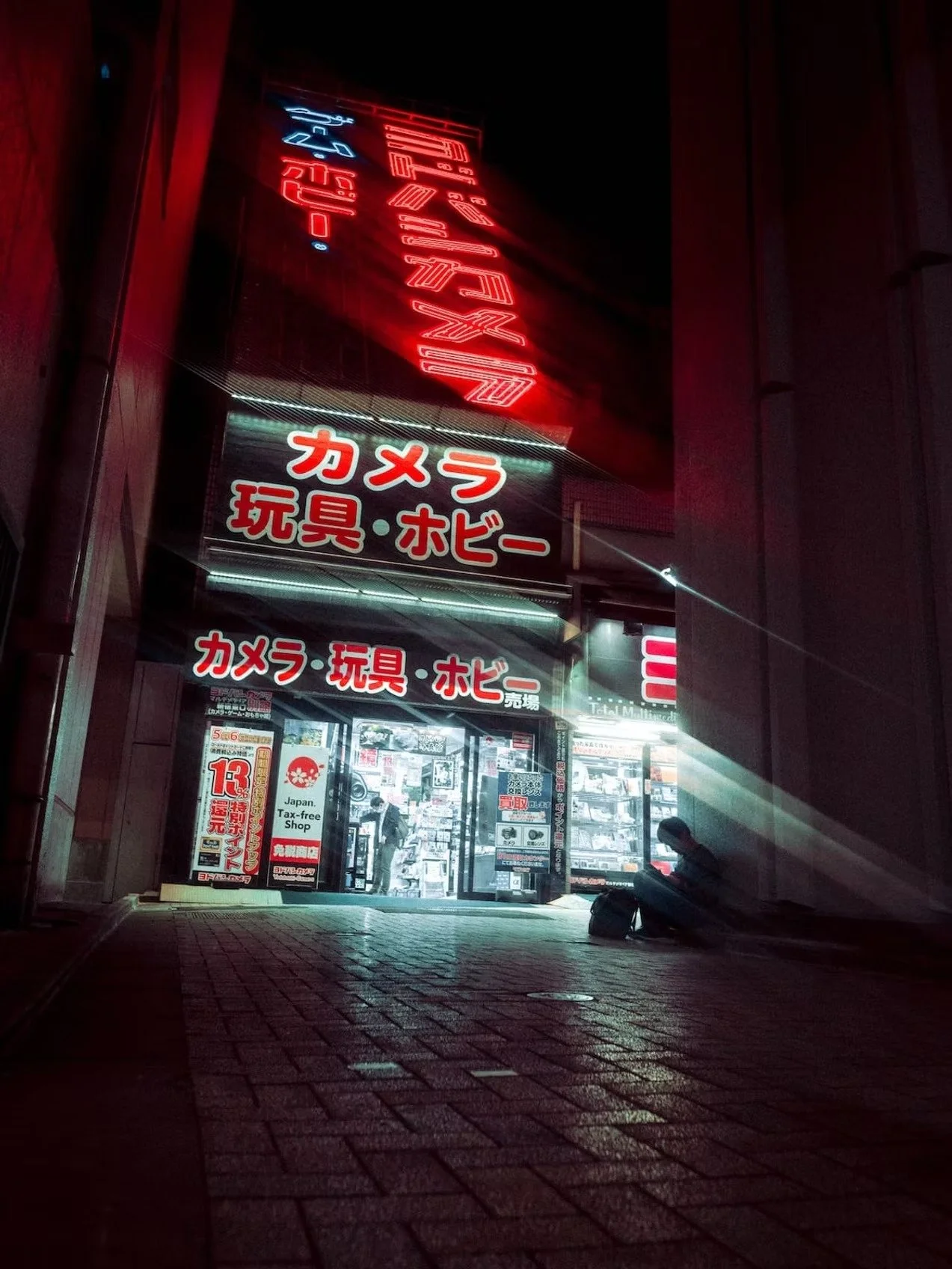Nighttime exterior view of a Japanese shop with illuminated red and white signage in Japanese characters, displaying toys, hobby items, and cameras, with a person sitting outside and a scooter on the street.